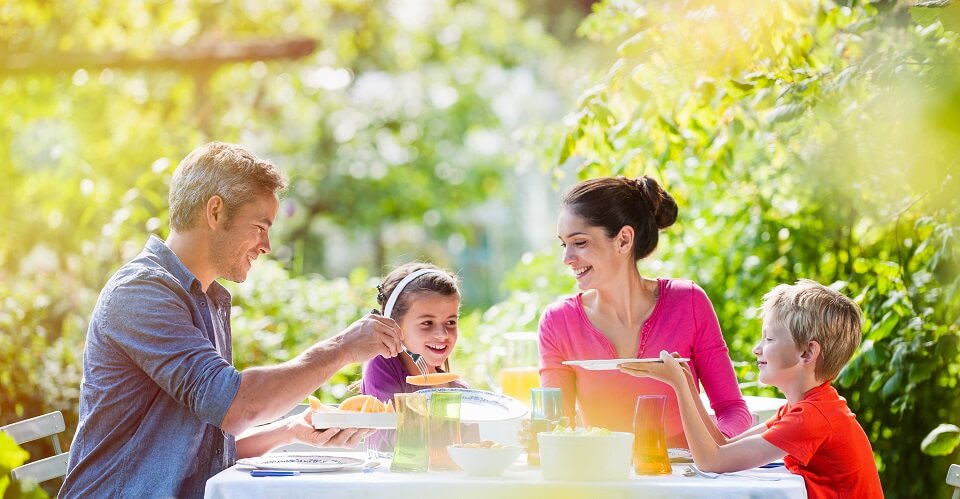 A man and a woman and a girl and a boy eat at a dining table with a white tablecloth in the yard